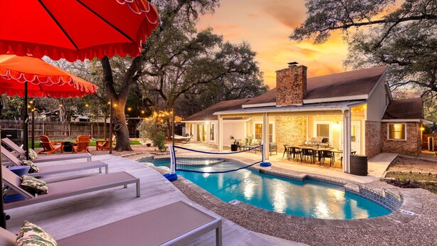 A pool area with lounge chairs, a red umbrella, and a building with a brick facade. - Ultimate Getaway • Pool • Game Room • Sleeps 12 (San Antonio)