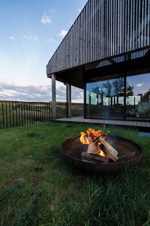 A modern outdoor fire pit area with a metal bowl, surrounded by grass, and a wooden deck leading to a glass-enclosed patio with outdoor furniture.