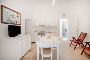 Una cucina moderna con mobili bianchi, un tavolo da pranzo con sedie, una televisione su un mobile e un quadro appeso al muro.