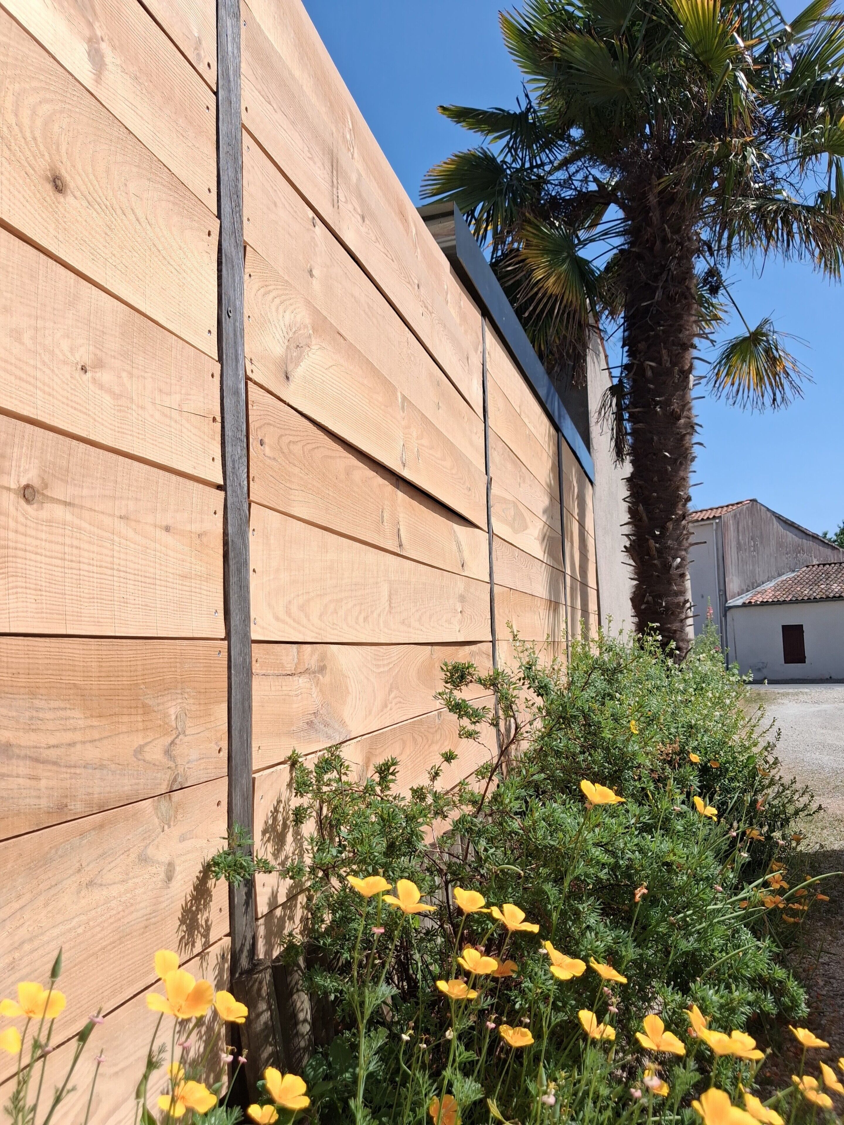 Pared de madera con un travesaño metálico vertical, una palmera y flores amarillas.