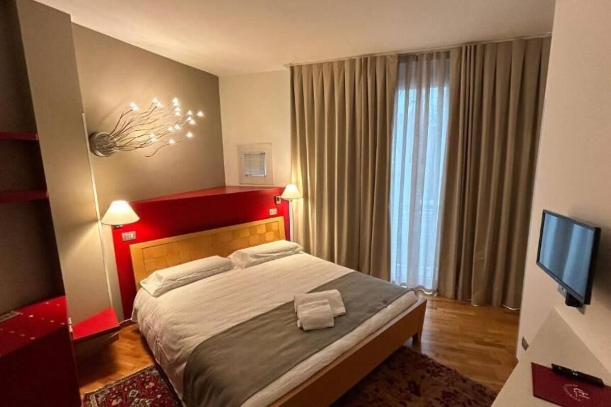 Ein Hotelzimmer mit einem großen Bett, einem an der Wand befestigten Fernseher, einem roten Kopfteil und einem Fenster mit Vorhängen.