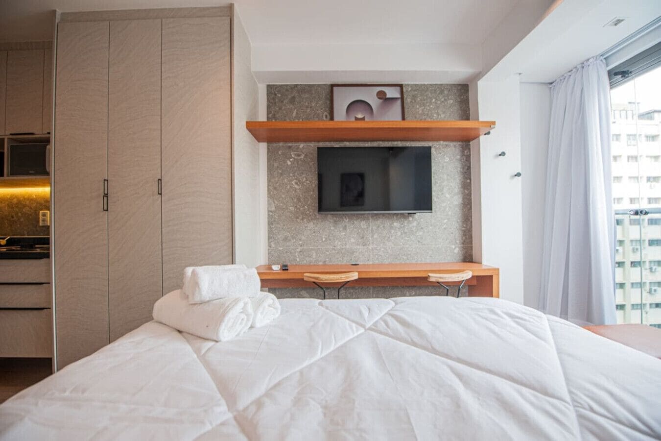 A modern hotel room with a large bed, a wooden desk, a flat-screen TV mounted on a textured wall, and a view of the city through a large window.