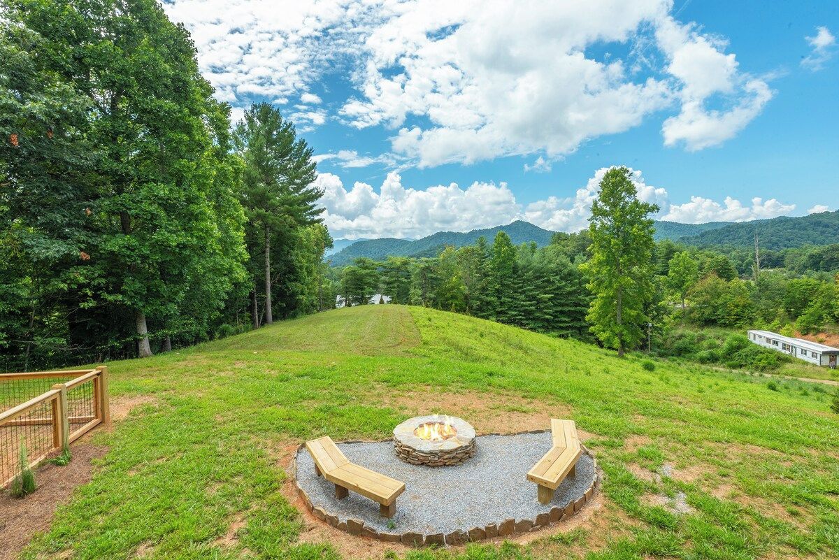 A circular outdoor seating area with a fire pit, surrounded by a wooden fence, set against a backdrop of a grassy hill and trees under a partly cloudy sky.