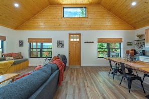 A spacious room with a high wooden ceiling, a large window, a cozy seating area, and a dining table with chairs. - Dreamcatcher Cabin~Hot Tub, Mtn Views & Fire Pit! (Burnsville)