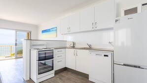 Uma cozinha moderna com armários brancos, forno embutido, máquina de lavar louça e frigorífico. Há uma janela com vista para o mar.