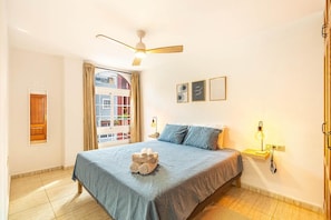 A bedroom with a bed, a ceiling fan, a window with curtains, a bedside table with a lamp, and a wall with framed pictures. - Vacation Apartment 'Starfish San Juan' with Mountain Views, Balcony & Wi-Fi (Guía de Isora)