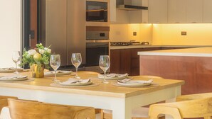 A dining area with a wooden table set for four, surrounded by wooden chairs with white cushions. A kitchen with modern appliances and a built-in oven is visible in the background.