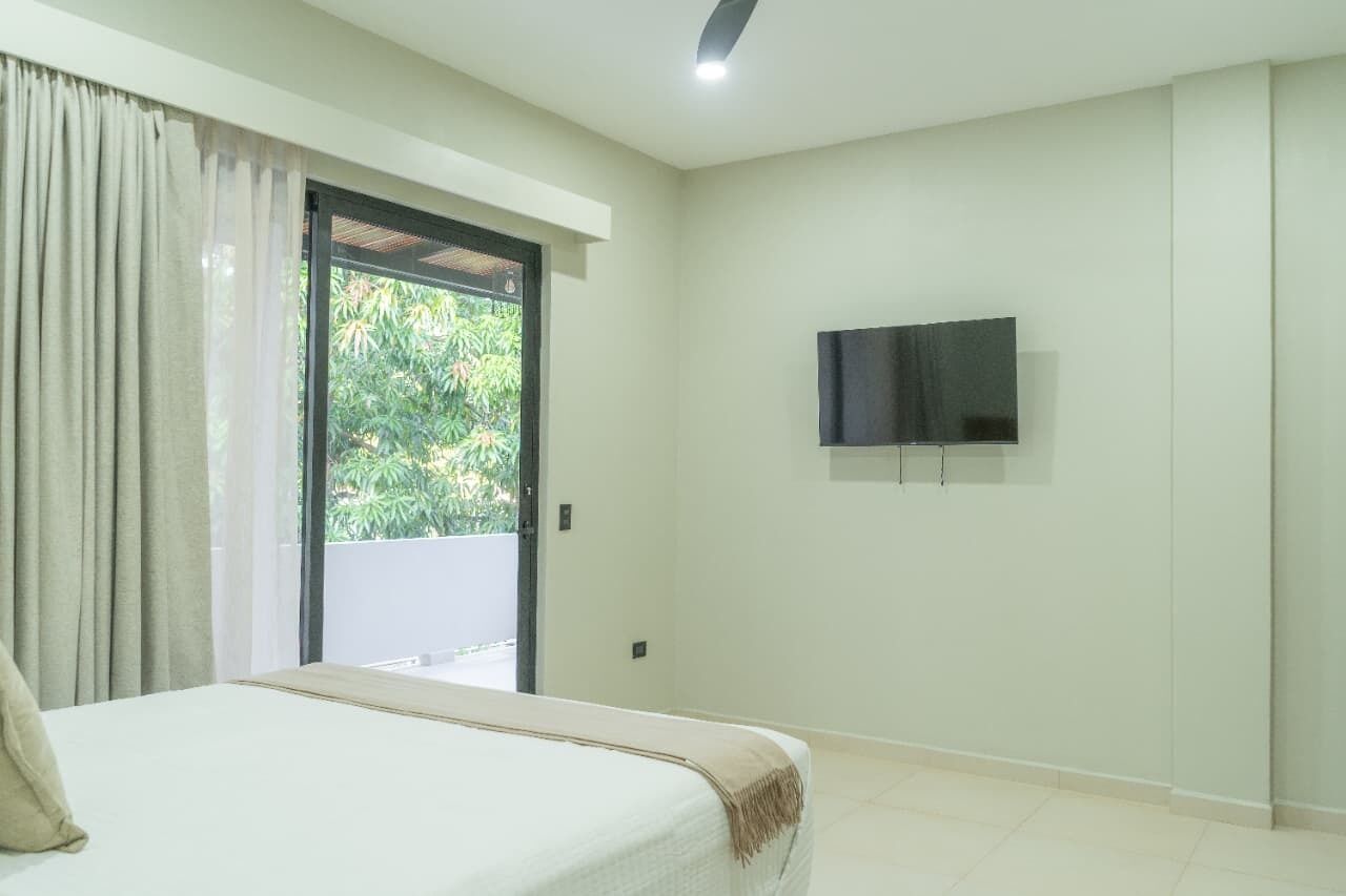A bedroom with a bed, a TV mounted on the wall, and a sliding glass door leading to a balcony.