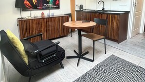 En kompakt opholdsstue med tekøkken, spiseplads med rundt bord og stole, sofa med gul pude og et tv ophængt på væggen.