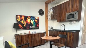 En moderne stue med et fladskærms TV, et træspisebord, en sort sofa og et køkken med træskabe og en mikroovn.