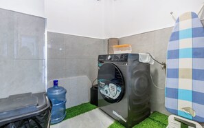 A compact laundry room with a front-loading washing machine, a large water dispenser, and a surfboard leaning against the wall. - VIP Duplex apartment with Coffe bar (Nairobi)