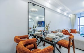 Un espace repas moderne avec une table en verre, des chaises recouvertes d’un tissu orange et un grand miroir qui reflète la pièce.