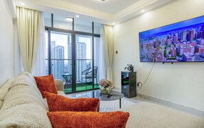 Una sala de estar moderna con un sofá, una mesa de centro, un televisor montado en la pared y vistas de un paisaje urbano a través de amplios ventanales.
