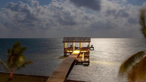 A wooden pier leading to a thatched-roof structure with seating, overlooking a calm sea under a cloudy sky. - Casa Al Mar Belize (St. George's Caye)