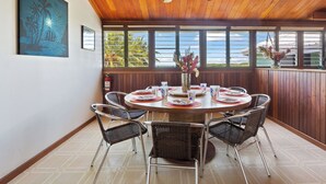 A dining area with a round table, wicker chairs, a ceiling fan, and a view of the ocean through large windows. - Casa Al Mar Belize (St. George's Caye)