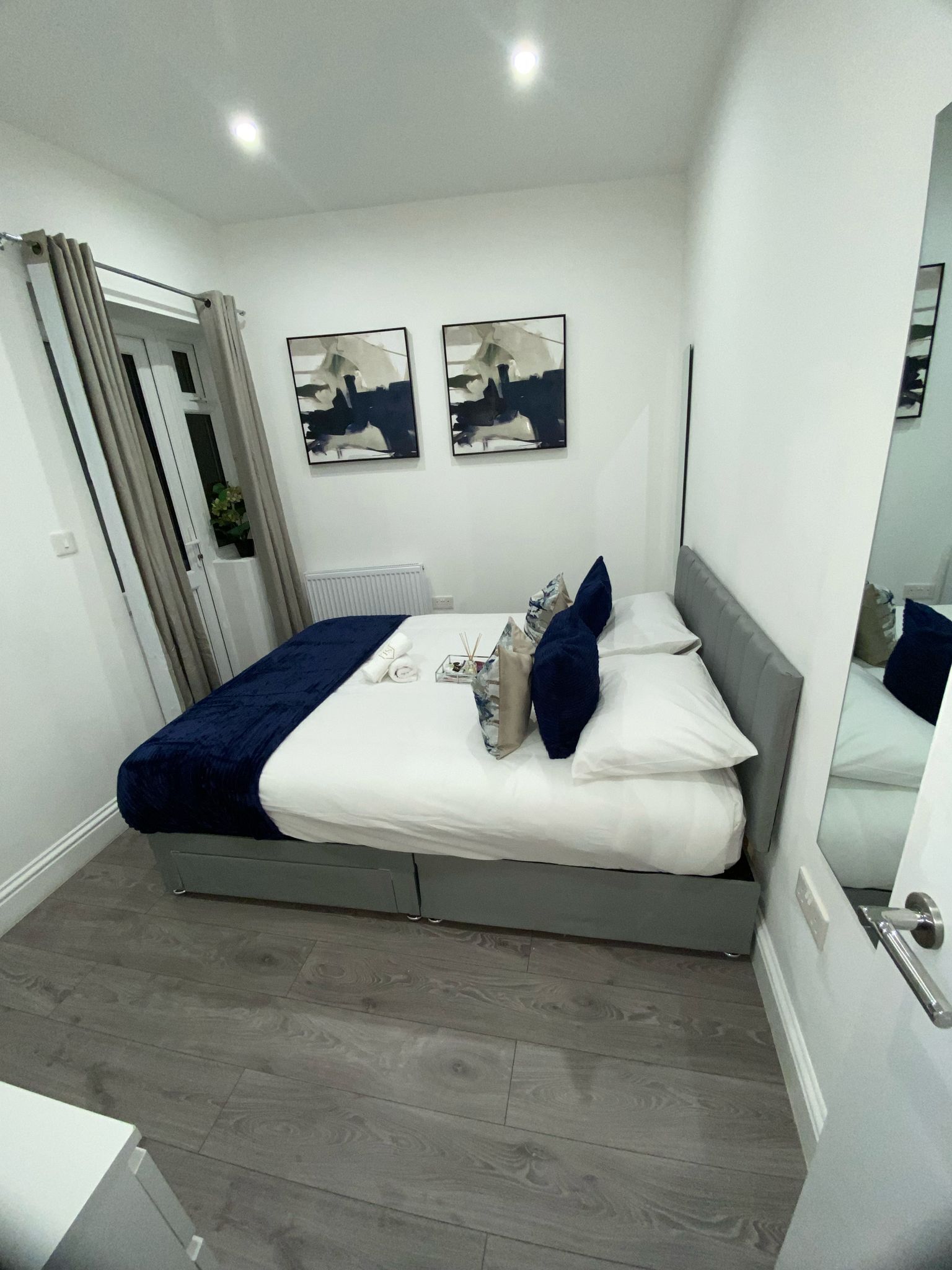 A modern bedroom with a bed, two wall-mounted abstract paintings, a radiator, and a mirror.