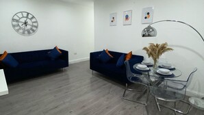 A modern living room with two blue sofas, a dining table with chairs, a wall clock, and abstract art. - Stylish 2bedroom Flat in London|free parking|Contractors (Wallington)