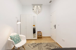 Una stanza con un'armadio bianco, uno sgabello di legno, una sedia bianca con cuscino decorato, una lampada da terra e un tappeto a motivi.