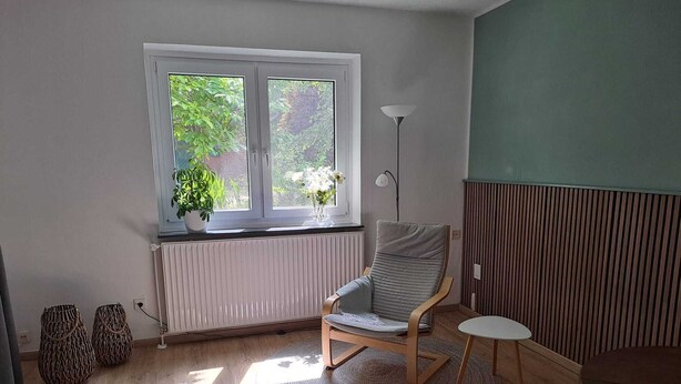 Ett rum med ett fönster, en element, en stol, ett bord och en golvlampa.