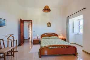 Un dormitorio con cama de madera, lámparas de noche, una silla de madera, una mesita, un cuadro en la pared y una ventana con cortinas.