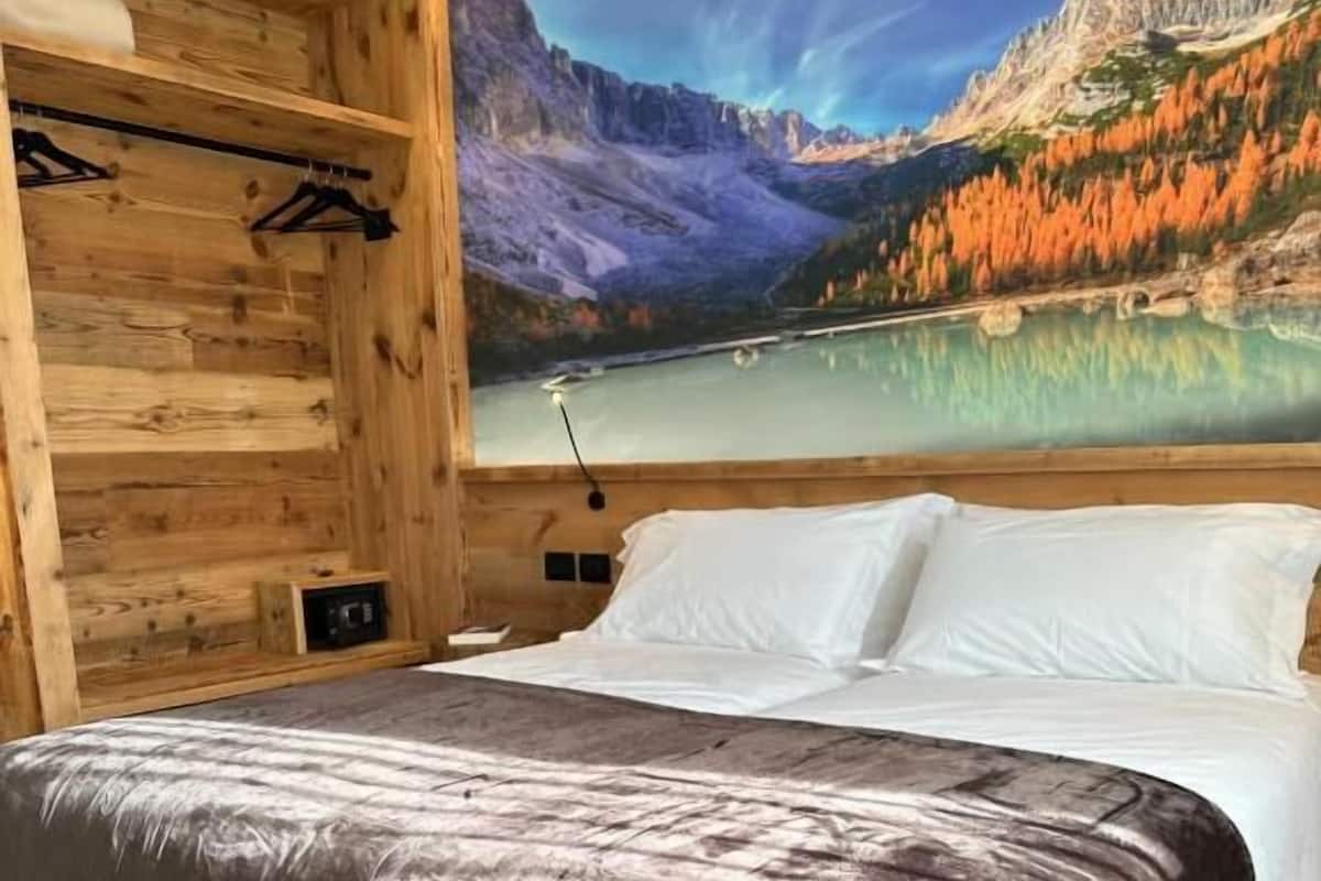 A bedroom with a wooden bed frame, a large bed with white linens, and a mural of a mountain landscape on the wall.