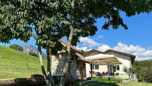 A stone house with a large umbrella patio, surrounded by a grassy hill and trees. - ORIGINI HOLIDAYS  (Sedico)
