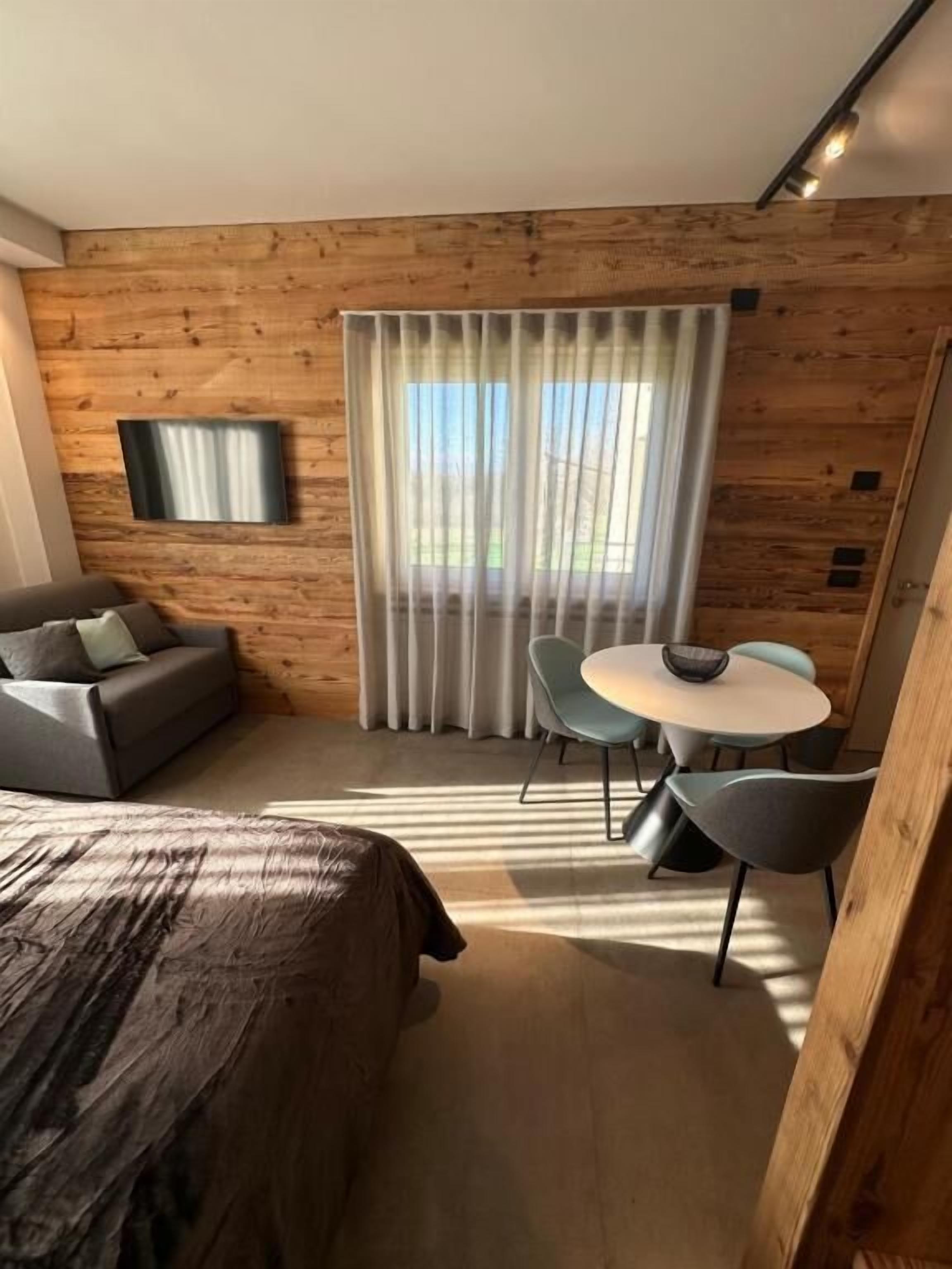 A modern room with a wooden wall, a bed, a TV, a dining table with chairs, and a window with curtains.