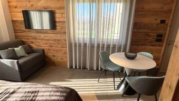 A modern room with a wooden wall, a bed, a TV, a dining table with chairs, and a window with curtains.