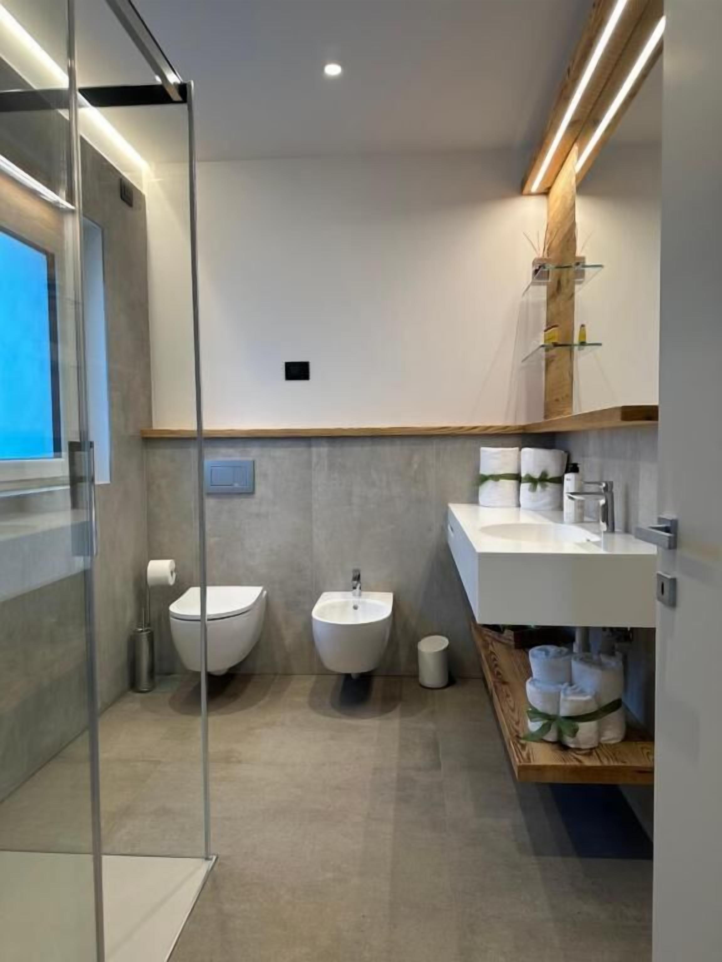 Ein modernes Badezimmer mit einer Duschkabine aus Glas, einem Bidet, einem Waschbecken und einem Holzboden mit Handtüchern.