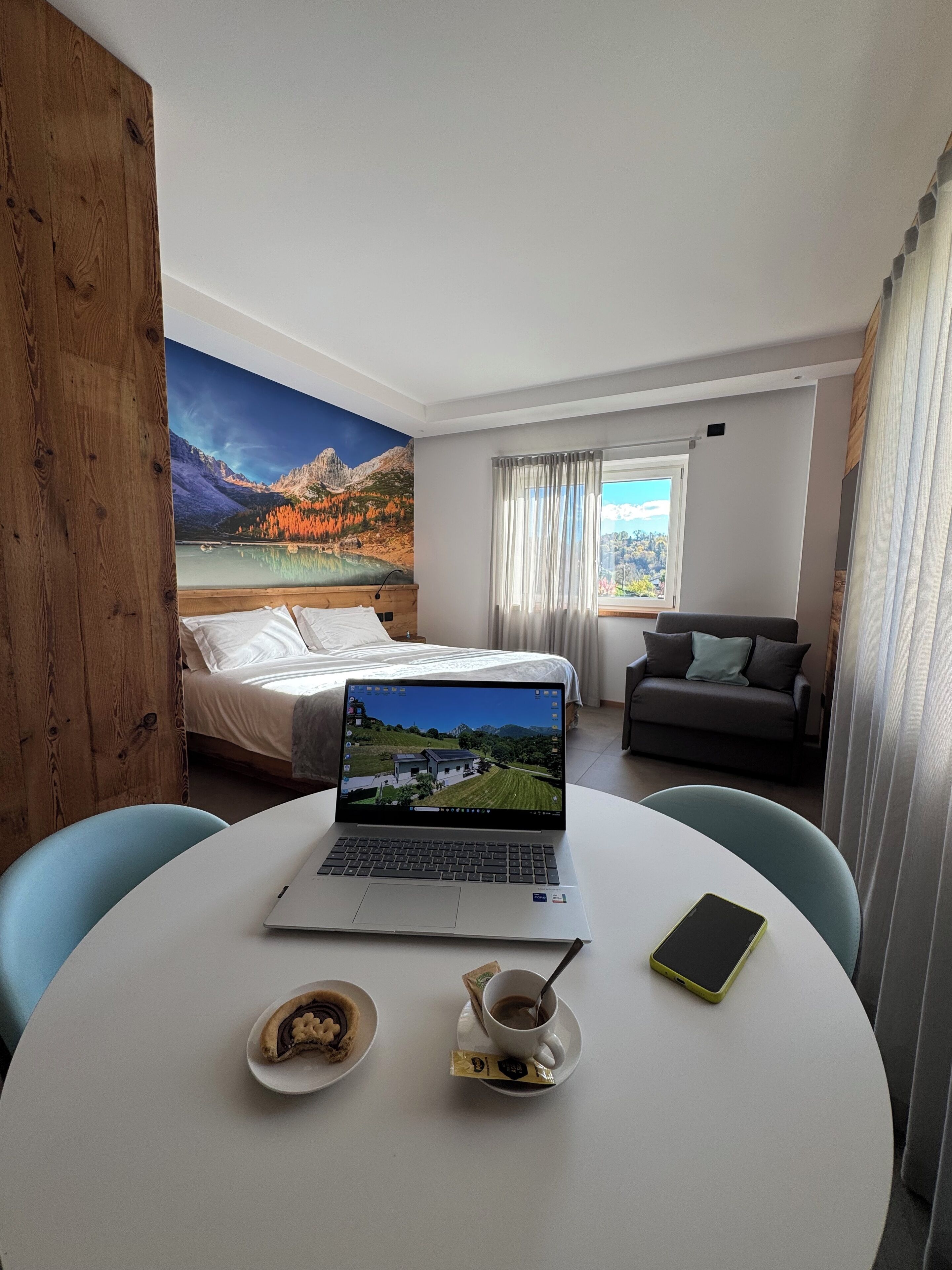 Camera d'albergo con un letto, un portatile su un tavolino rotondo, un divano e vista sulle montagne dalla finestra.