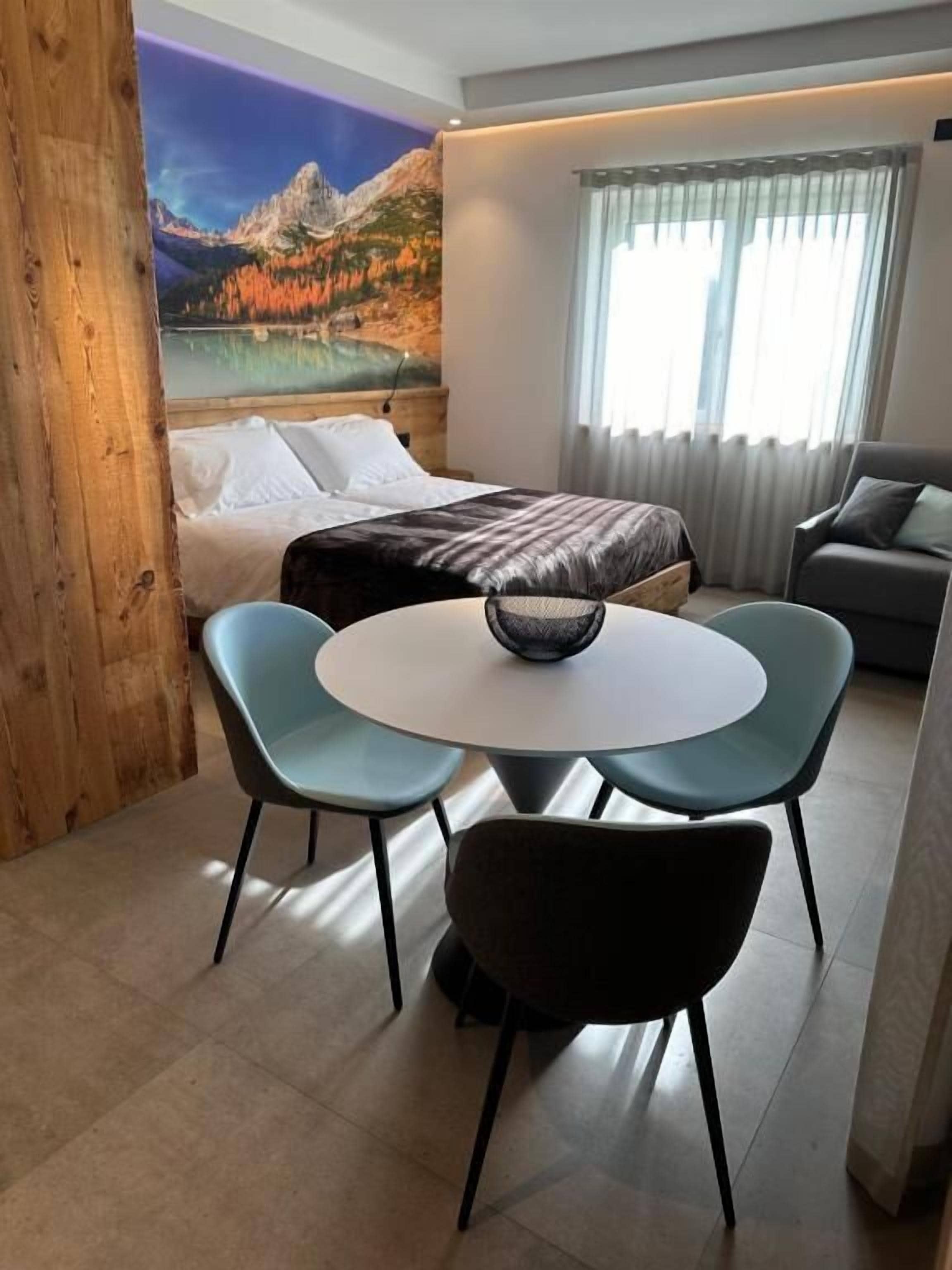 Una habitación moderna con cama, una mesa redonda con sillas, un sofá y un cuadro grande de un paisaje montañoso.