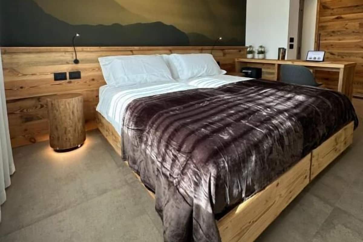 A modern bedroom with a wooden headboard, a large bed with a dark patterned bedspread, a nightstand with a lamp, and a wall-mounted TV.