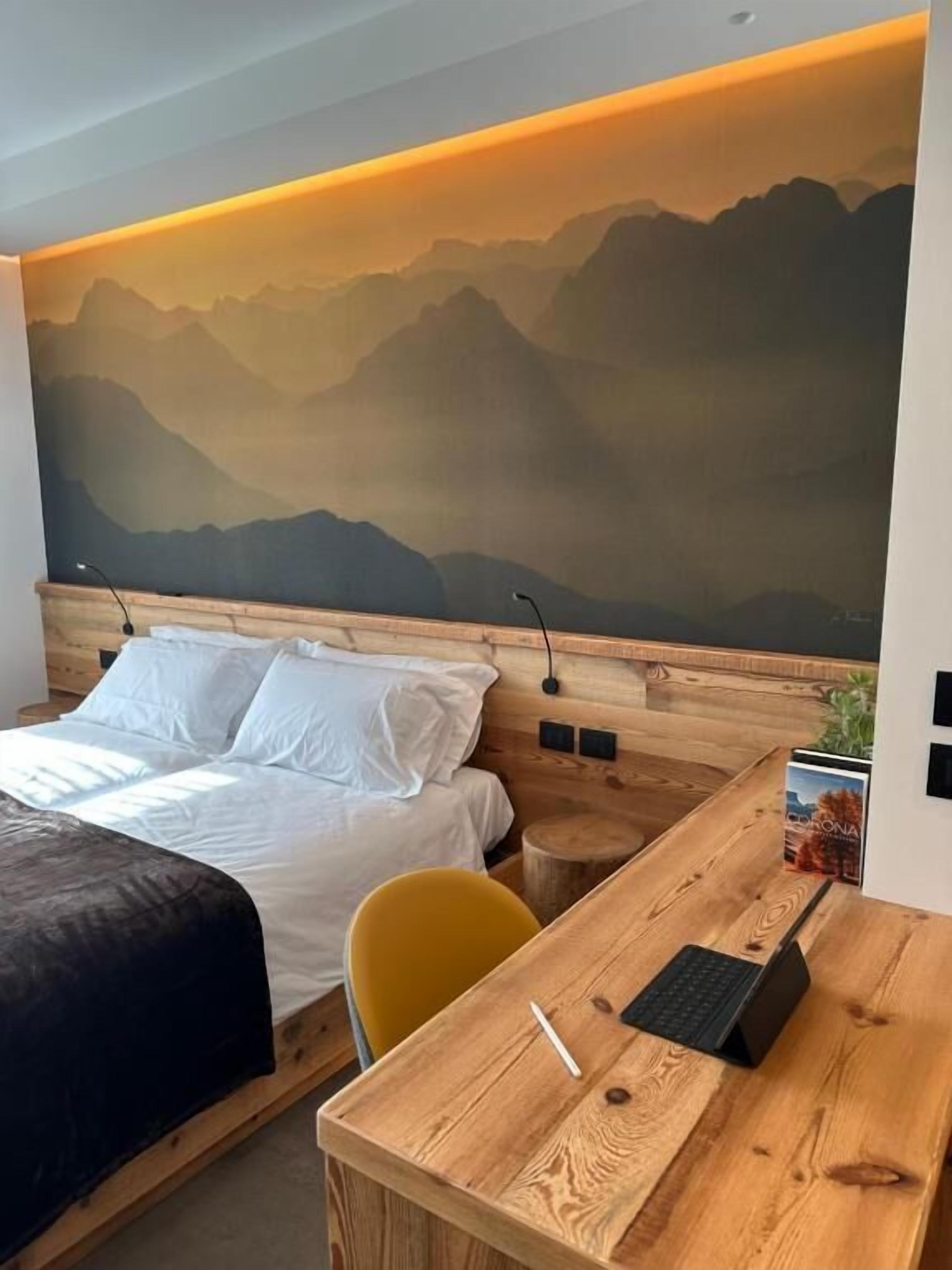 A bedroom with a bed, a desk with a laptop, a chair, and a wall mural of mountains.