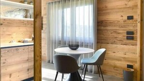 A modern interior with a wooden wall, a round table with chairs, and a window with curtains. - ORIGINI HOLIDAYS  (Sedico)