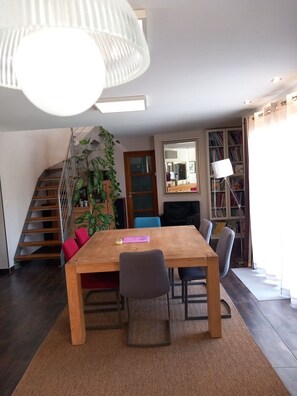 A dining area with a wooden table, chairs, a staircase, a bookshelf, and a hanging light fixture.