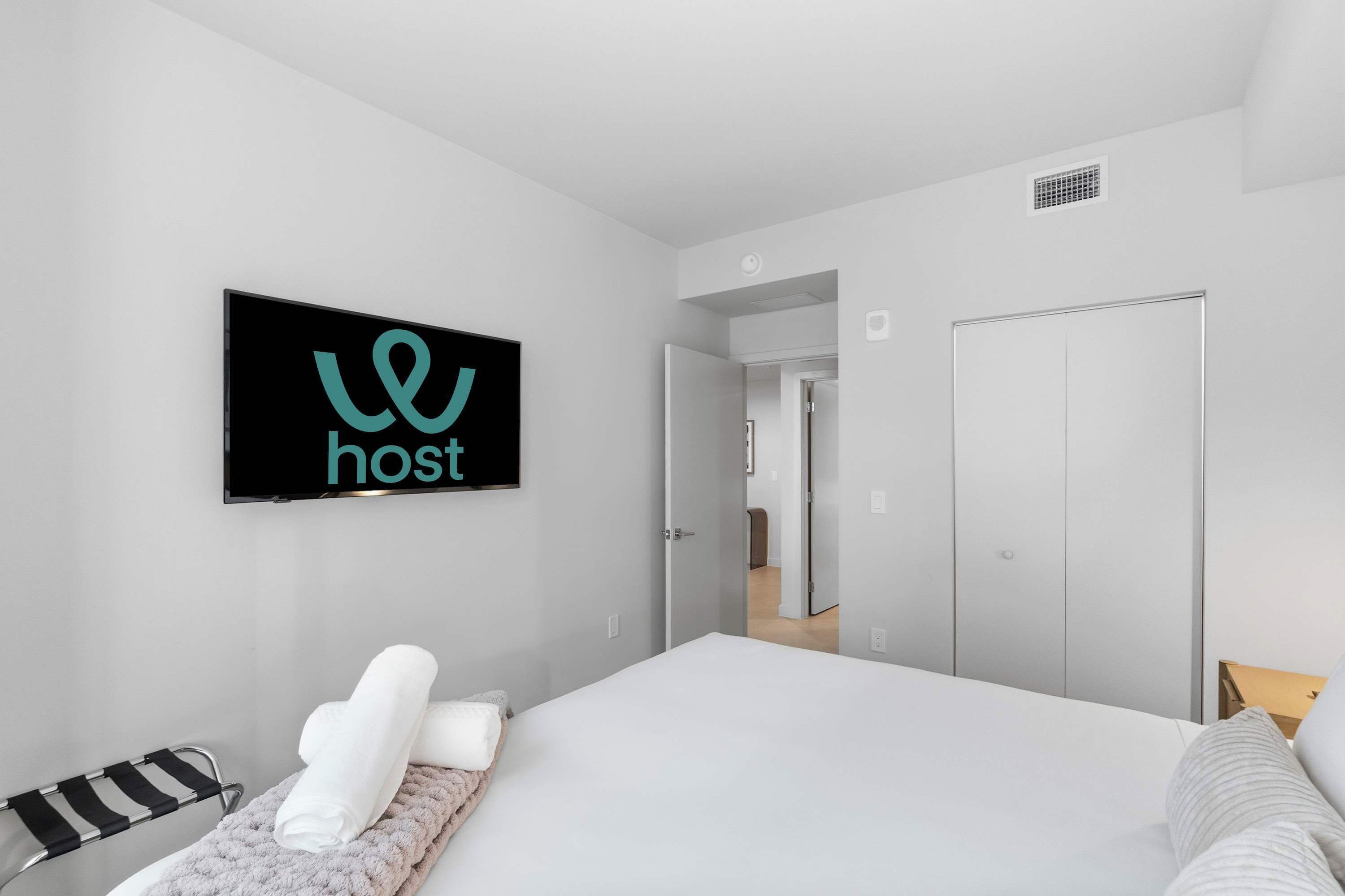 Une chambre d’hôtel moderne avec un lit, un téléviseur fixé au mur qui affiche un logo, une serviette sur le lit et une porte donnant sur une autre pièce.