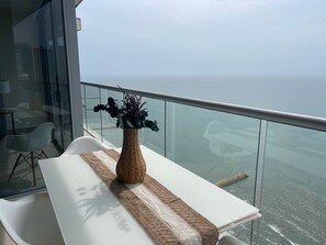 Un balcon avec une table blanche, un vase en osier rempli de fleurs et une vue sur l’océan.