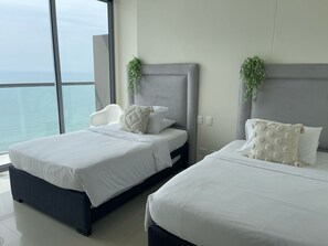 Une chambre d’hôtel avec deux lits, une grande fenêtre donnant sur la mer et une tête de lit grise.