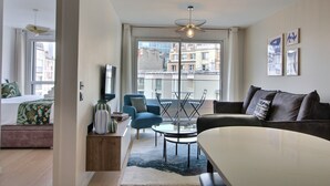 Una sala de estar moderna con un sofá, una mesa de centro y un sillón azul. Se aprecia una ventana grande con vistas a edificios.