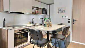 Una cocina compacta con horno integrado, microondas y un refrigerador pequeño. Hay una mesa de comedor con cuatro sillas y una bolsa de comestibles encima.