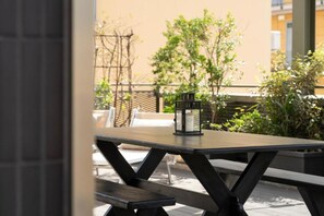 Un patio al aire libre con una mesa y bancos de madera, un farol y vistas a un edificio y vegetación.