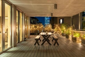 Un patio al aire libre con una mesa y bancos de madera, rodeado de plantas en macetas y luces colgantes.