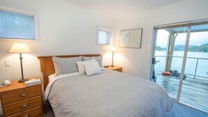 Een slaapkamer met een bed, nachtlampjes, een nachtkastje en uitzicht op een waterpartij door een schuifdeur.