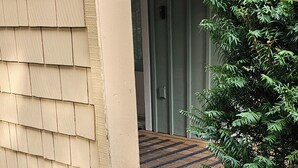 Een houten helling die naar een gebouwde ingang leidt, met een beige gevel en een klein groene plantje ernaast.