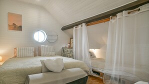 Une chambre avec un plafond en pente, une fenêtre ronde, un lit avec une literie blanche, un banc et une table de chevet avec une lampe.