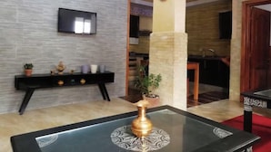 A modern interior with a flat-screen TV mounted on a textured wall, a console table with decorative items, a glass coffee table with a decorative object, and a kitchen area visible through an open doorway. - Apartment Youssef 5 min walk from the beach (Essaouira)