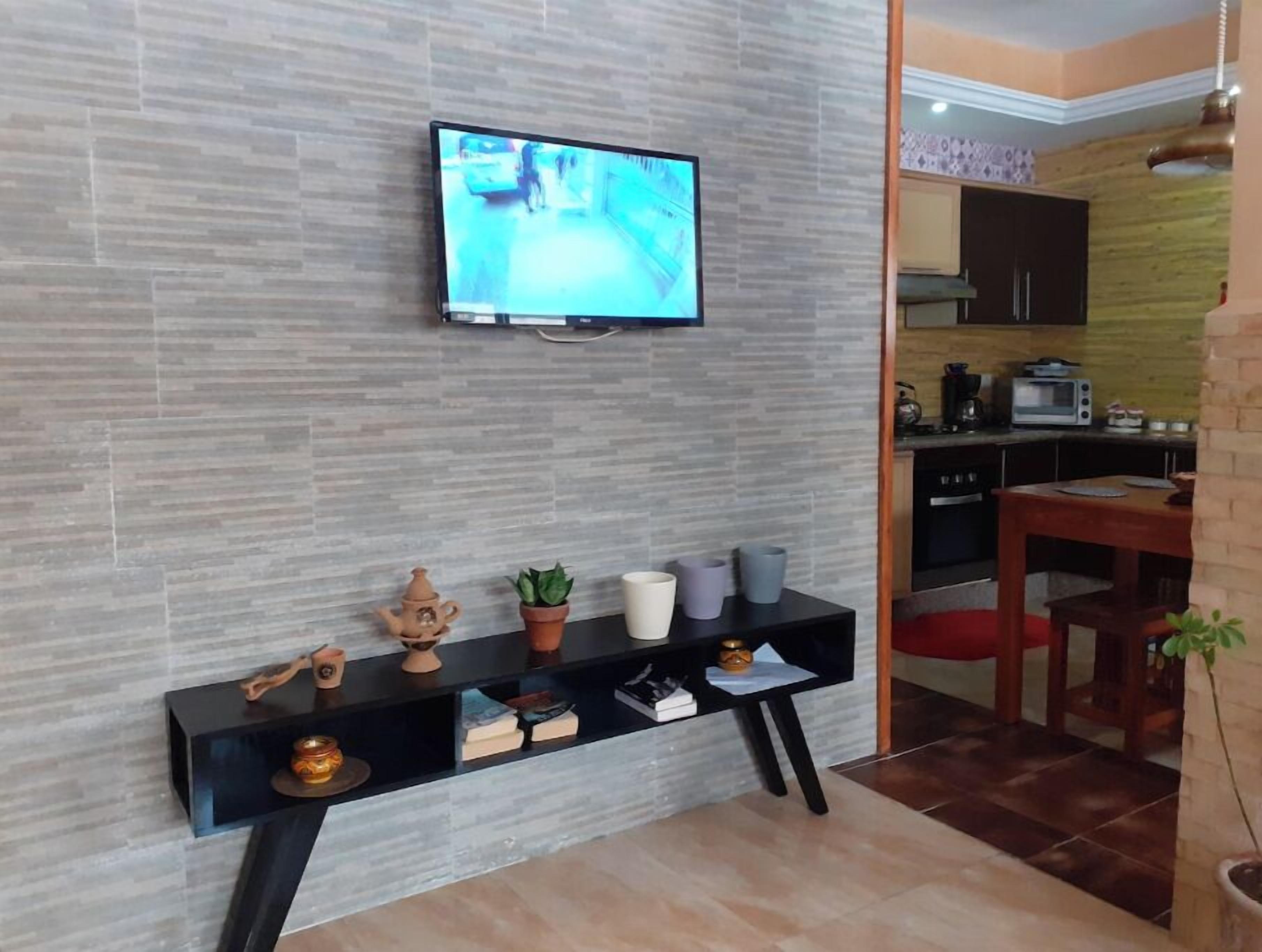 A modern living room with a flat-screen TV mounted on a textured wall, a black console table with decorative items, and a kitchen area visible through an open doorway.