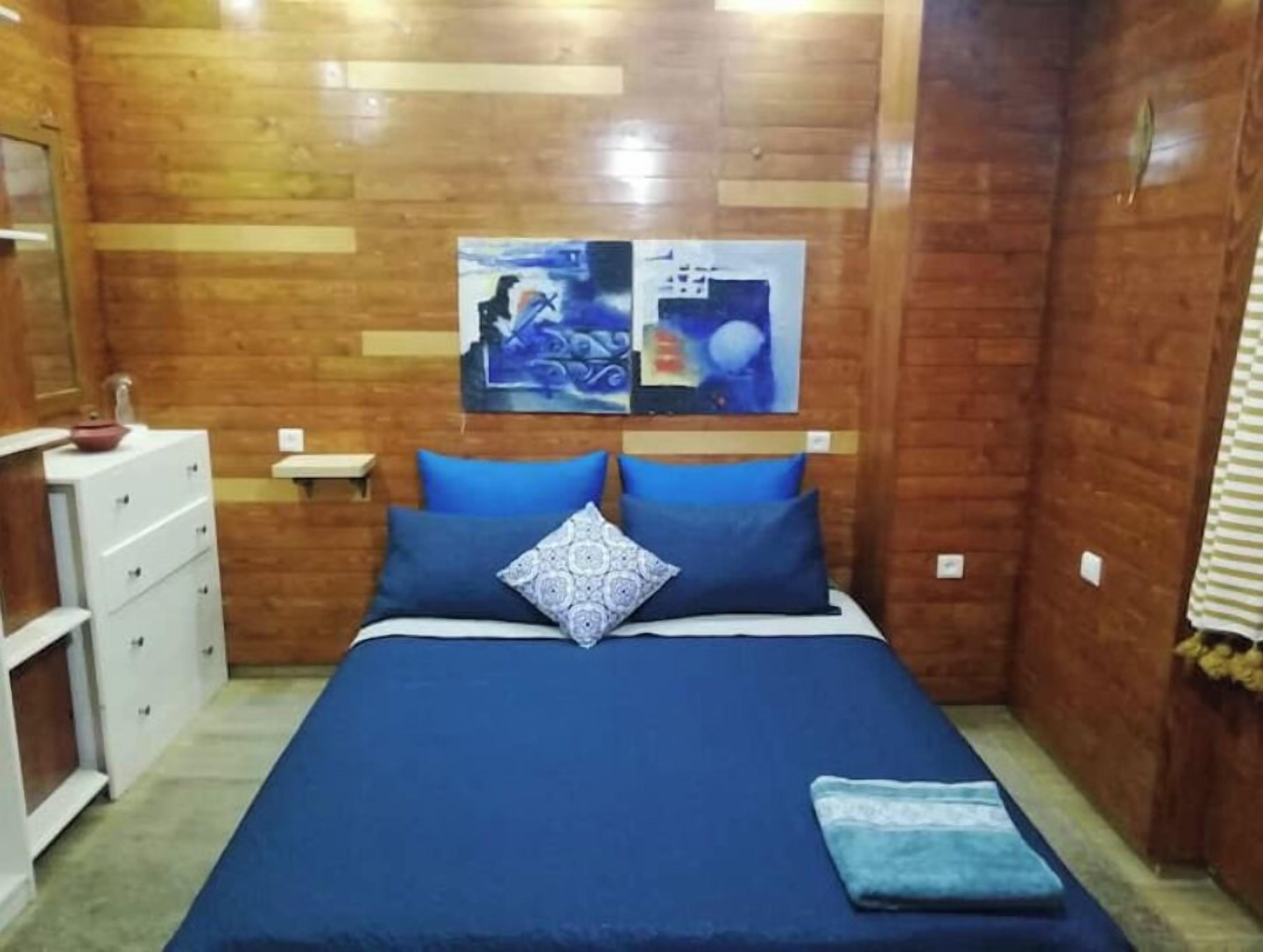 Une petite pièce chaleureuse avec un mur en bois, un lit avec une literie bleue, une commode blanche et une petite table.