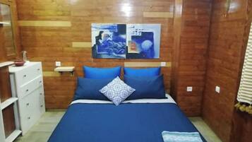 Une petite pièce chaleureuse avec un mur en bois, un lit avec une literie bleue, une commode blanche et une petite table.