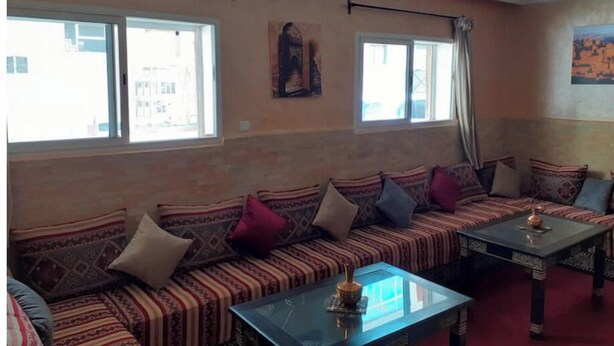 A room with a long sofa set, a glass-top table, and a window with a view of buildings. - Apartment Youssef 5 min walk from the beach (Essaouira)